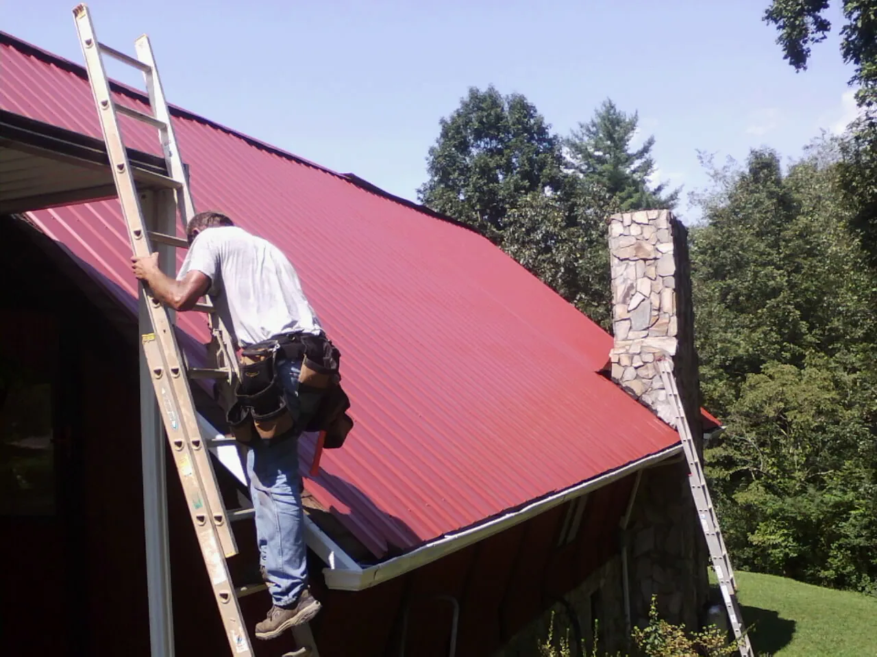 Roofer on a ladder beside a red metal roof and stone chimney — Soffit & Fascia Repair by Roofing Brothers in High Falls